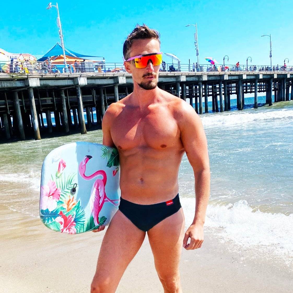 Man wearing black swim briefs holding a colorful bodyboard on the beach.